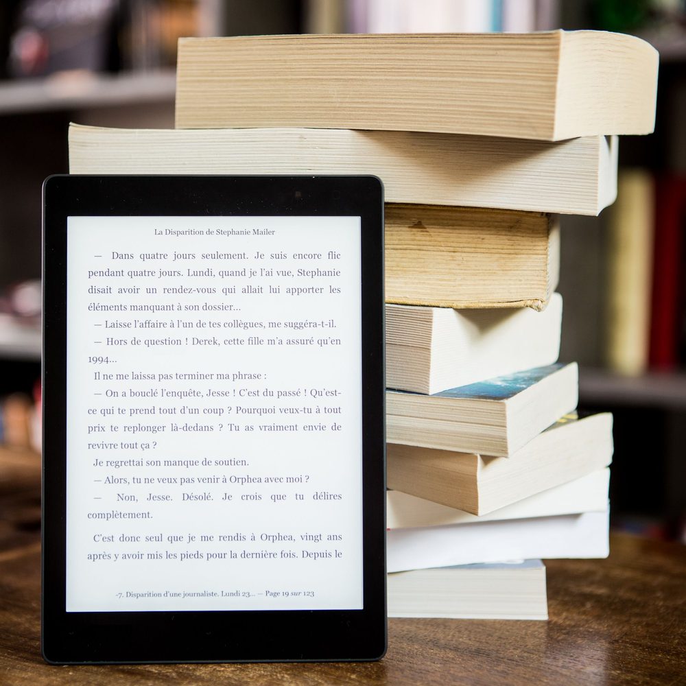 eReader mit aufgeschlagener eBook-Seite vor einem Stapel Bücher auf einem Holztisch